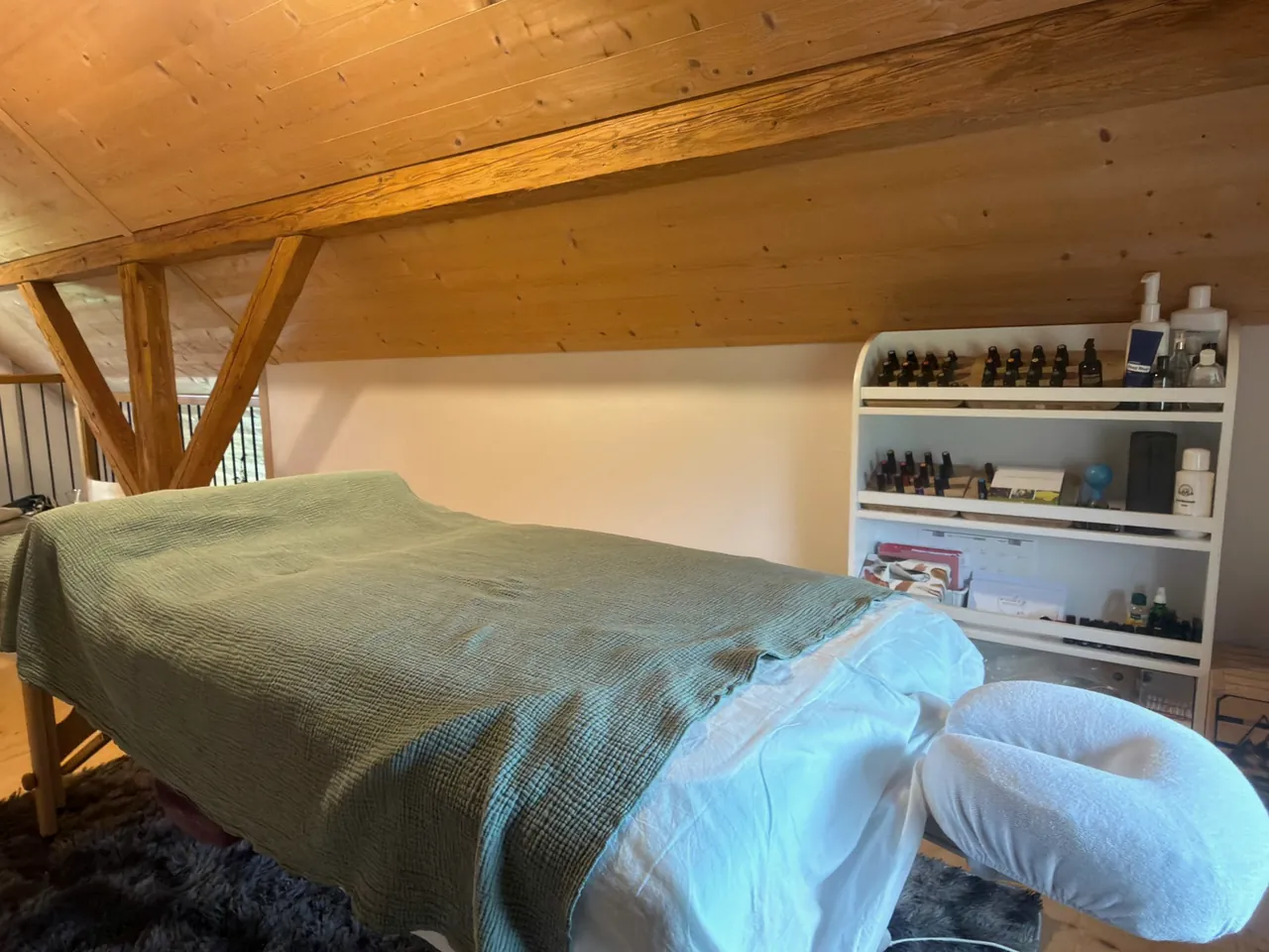 Behandlungsraum mit Massageliege unter dem Dachgebälk und einem Regal mit ätherischen Ölen