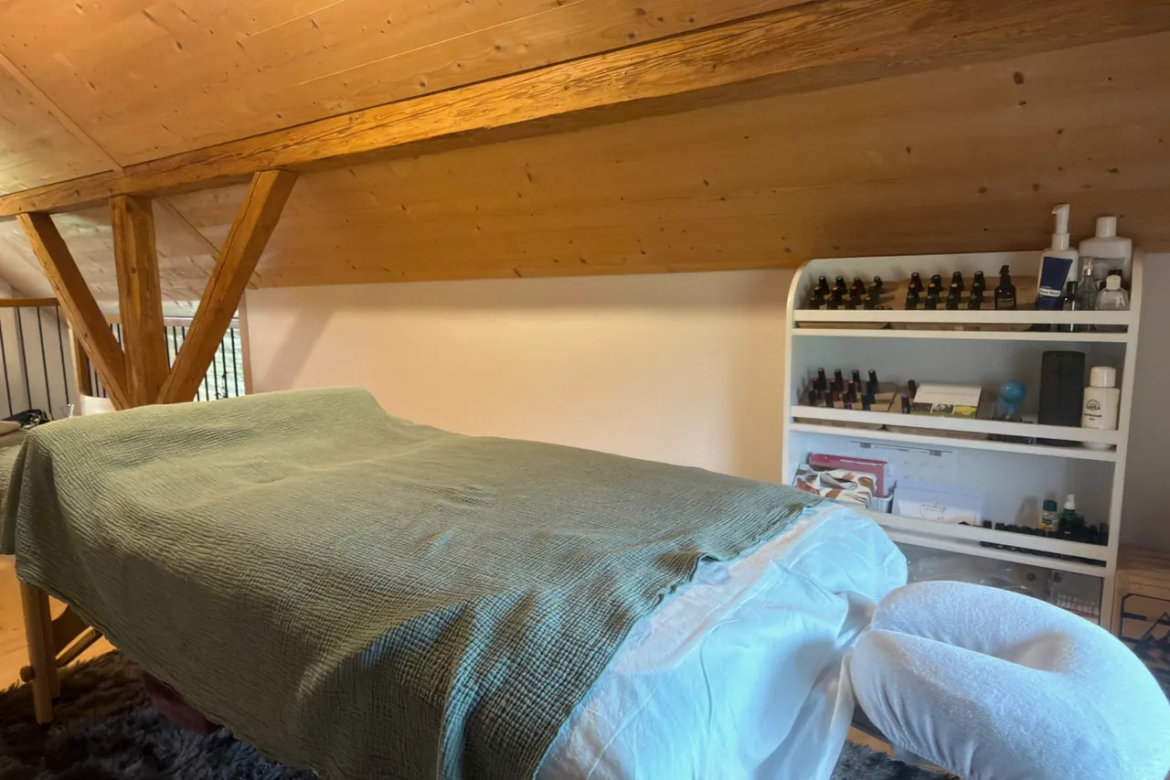 Behandlungsraum mit Massageliege unter dem Dachgebälk und einem Regal mit ätherischen Ölen